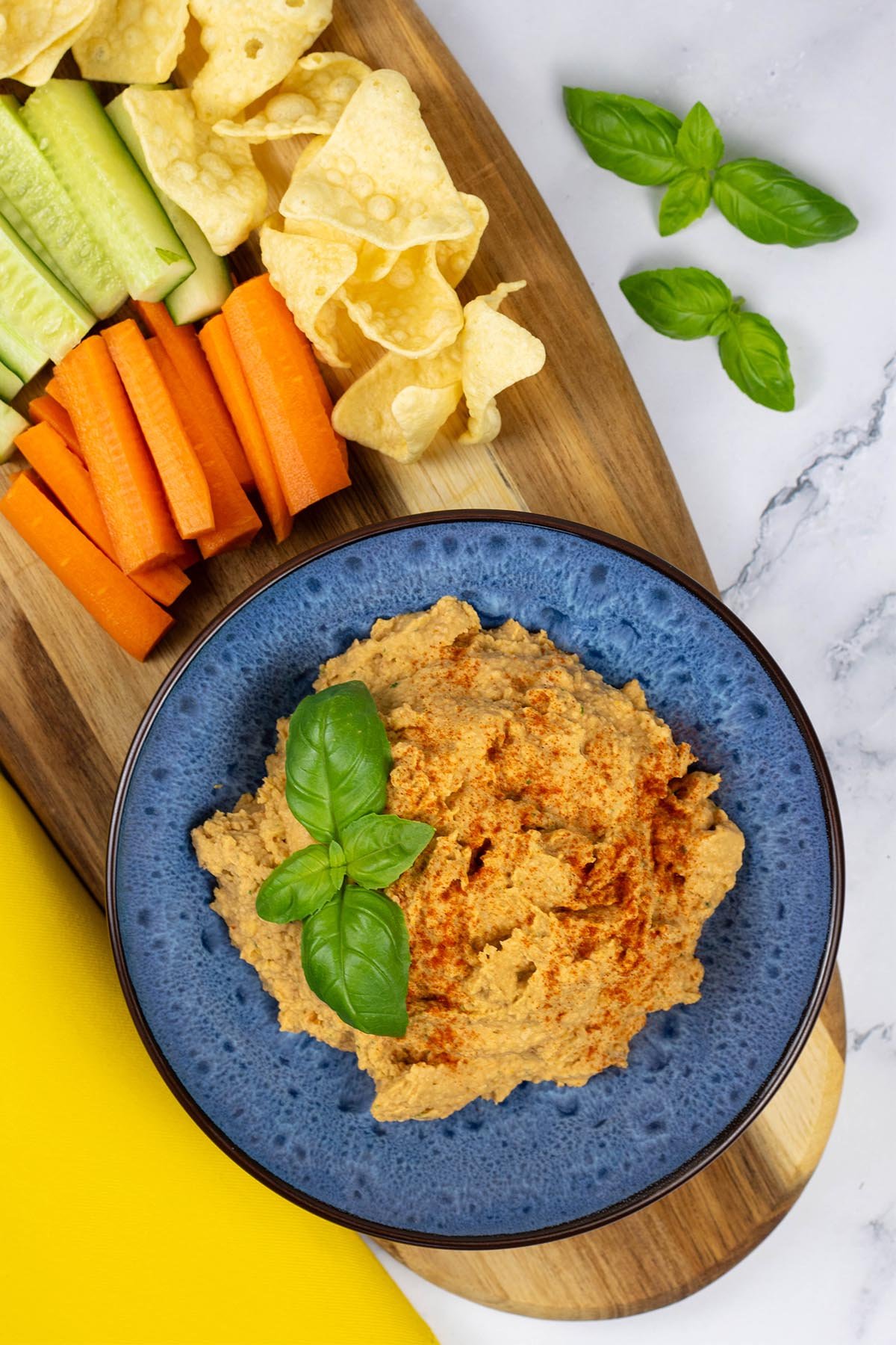 Sun-dried tomato and basil houmous in a blue patterned serving bowl sitting on a board with carrot and cucumber sticks and lentil crisps.