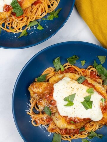 2 bowls of turkey parmigiana with spaghetti in blue bowls with a mustard napkin on the side.