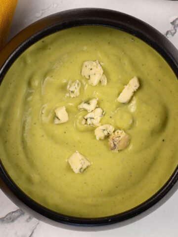 A portion of broccoli and stilton soup garnished with crumbled stilton in a black bowl, made in the Thermomix.
