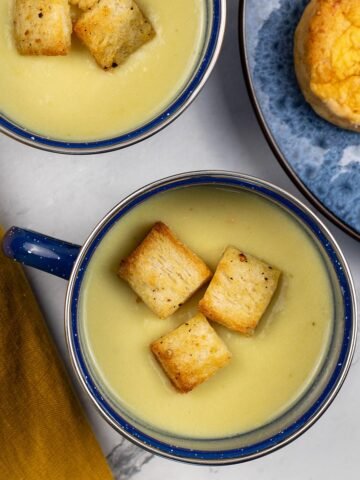 Two portions of cauliflower cheese soup in blue enamel mugs with a plate of chilli cheese scones on the side.