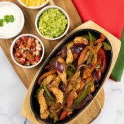 A portion of chicken fajitas in a sizzle platter sitting on a board with small bowls of sour cream, guacamole, grated cheese and pico de gallo salsa on the side.