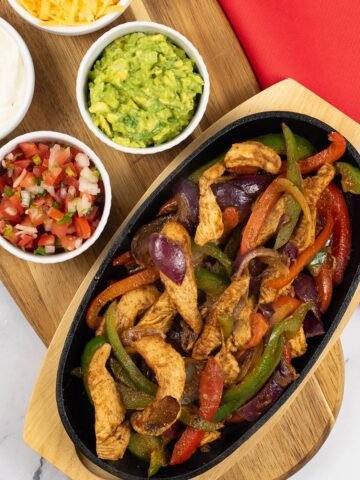 A portion of chicken fajitas in a sizzle platter sitting on a board with small bowls of sour cream, guacamole, grated cheese and pico de gallo salsa on the side.