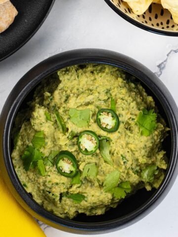 Coriander and green chilli houmous in a black serving bowl garnished with coriander leaves and sliced green chillis.