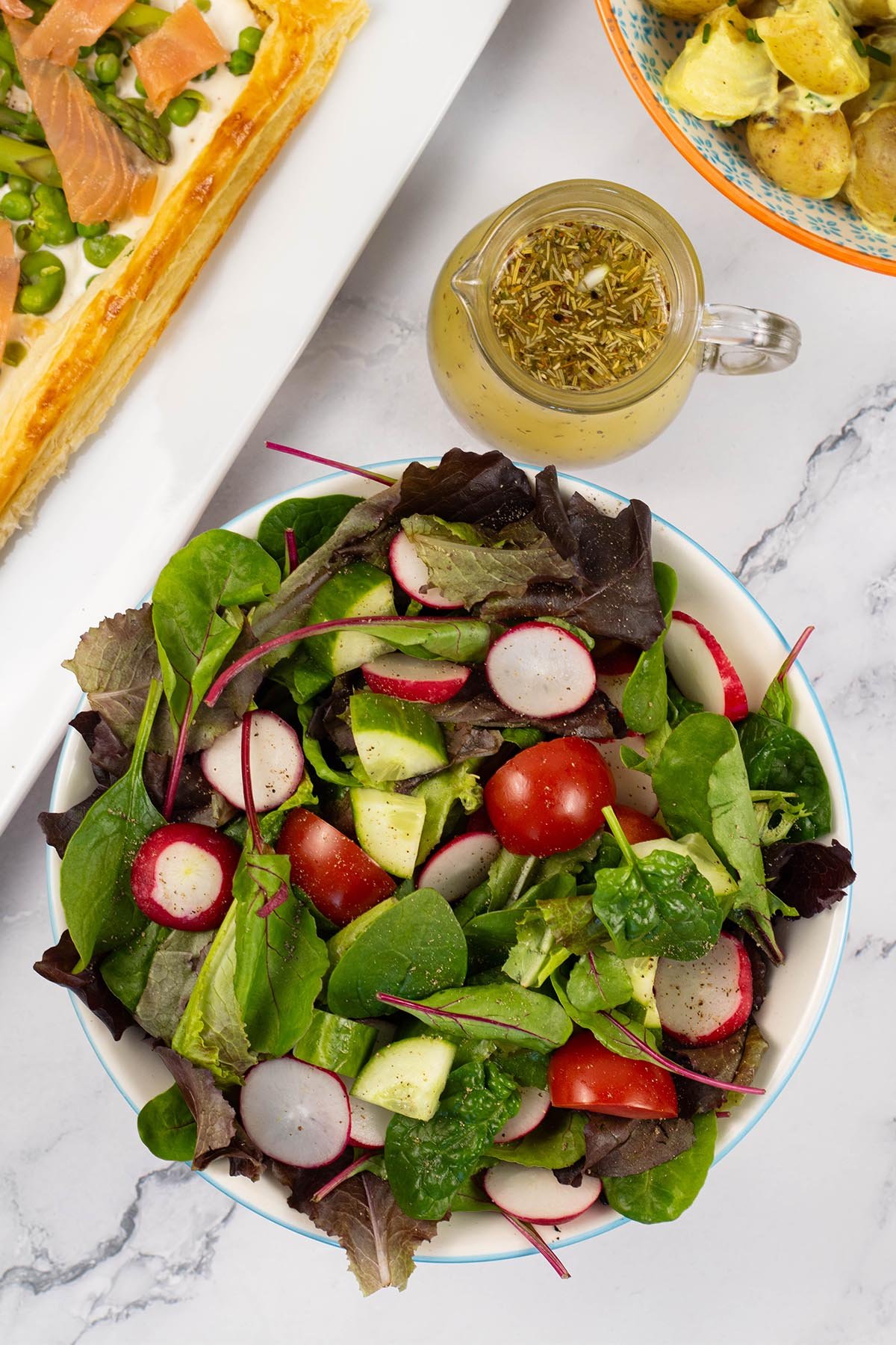 Easy baby leaf salad in a light blue rimmed serving bowl with a jug of zesty Italian-style dressing on the side.