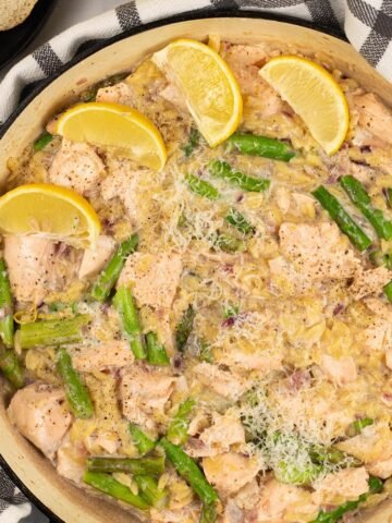 Salmon and asparagus orzo in a large round shallow casserole, garnished with parmesan and lemon wedges.