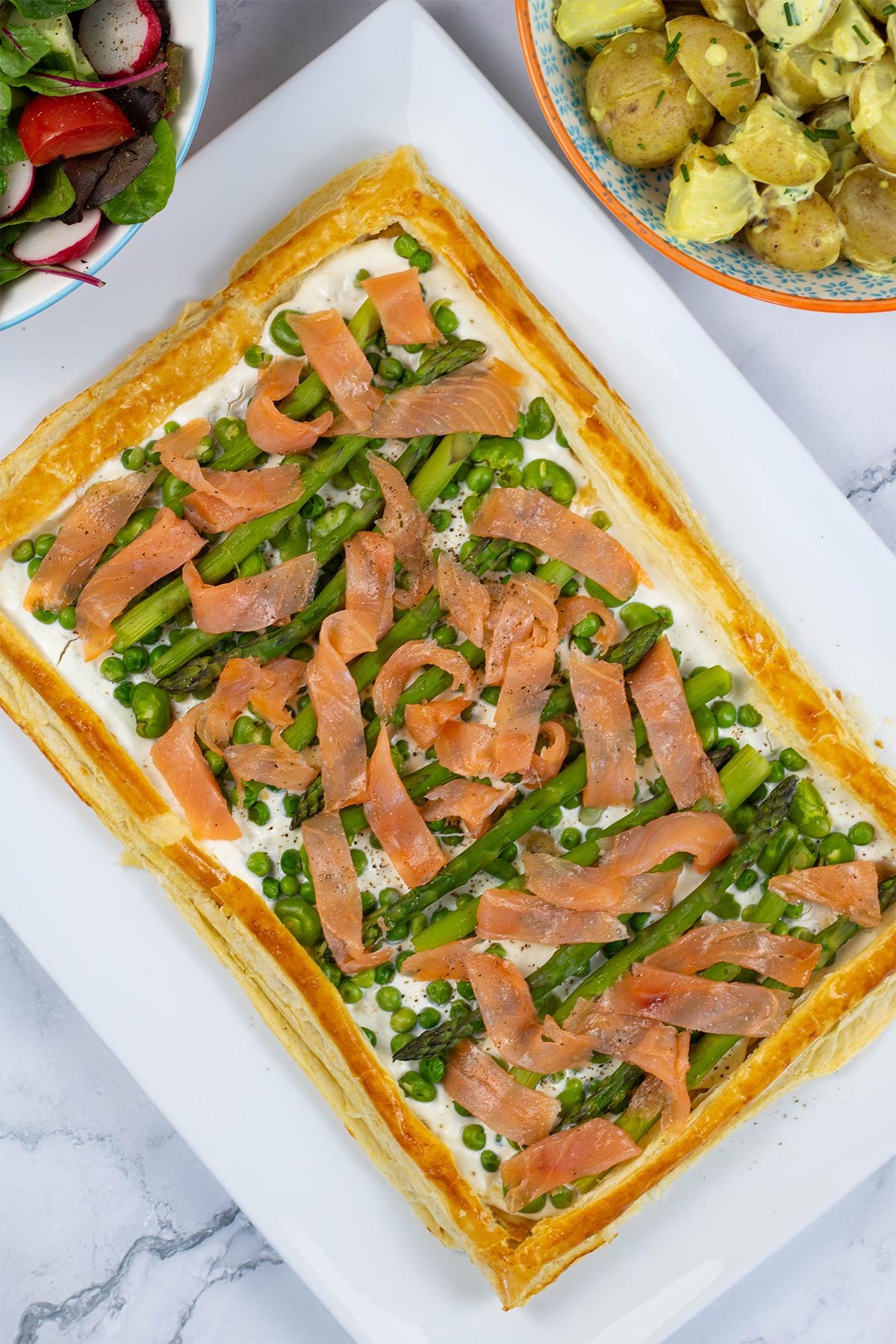 Smoked salmon and spring vegetable tart on a white rectangular serving platter, with bowls of potato salad and easy baby leaf salad on the side.