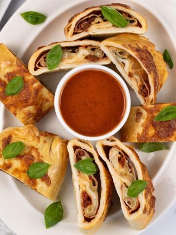Sliced stromboli on an oval white platter, garnished with basil leaves and served with a dish of tomato pizza sauce.