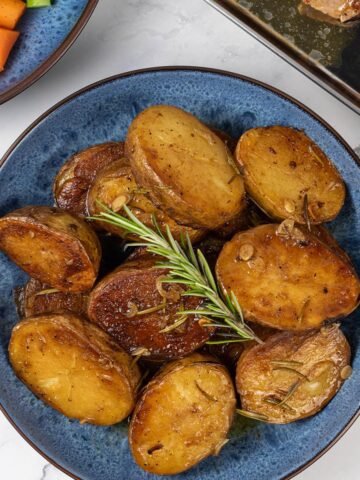 Buttery herbed melting potatoes in a blue patterned serving bowl, with a bowl of carrots and broccoli and tray of pork chops in the background.