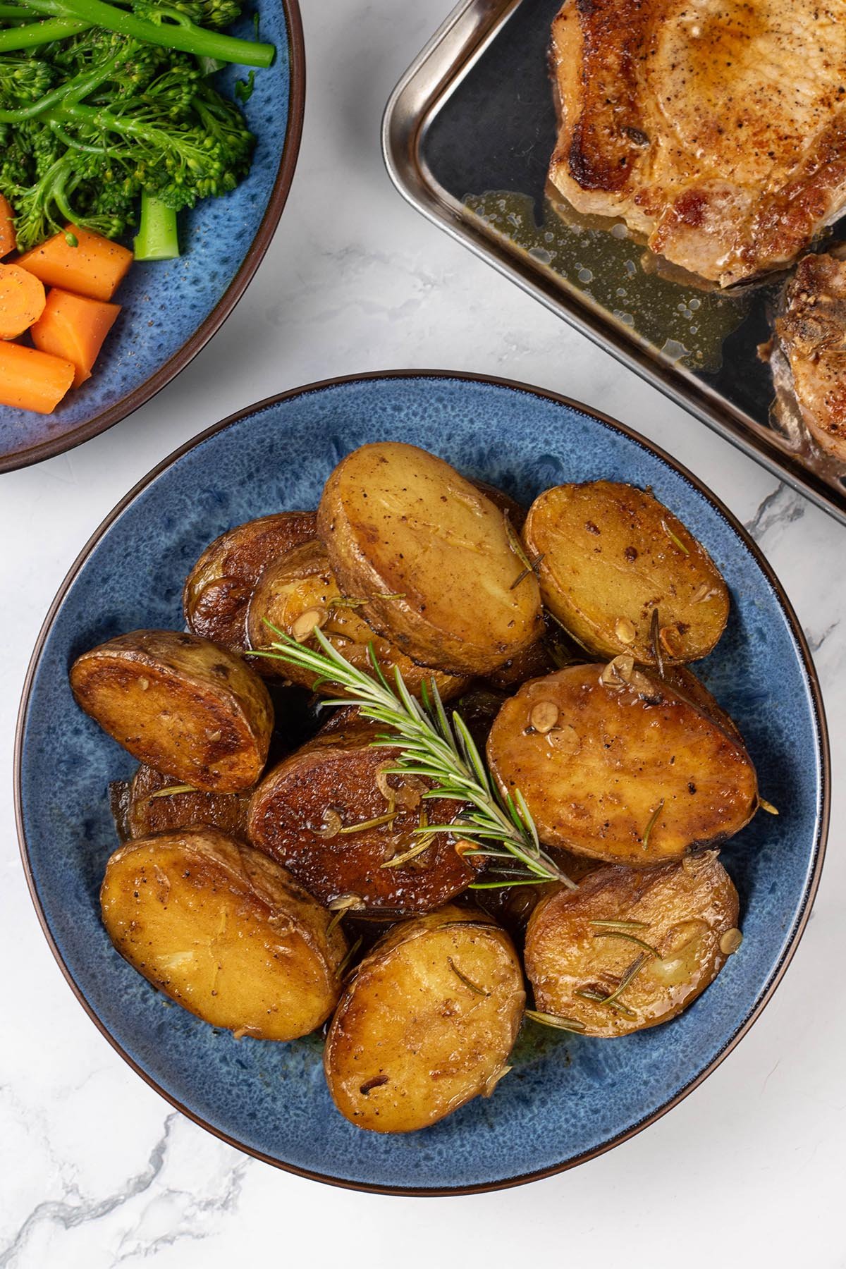 Buttery herbed melting potatoes in a blue patterned serving bowl, with a bowl of carrots and broccoli and tray of pork chops in the background.