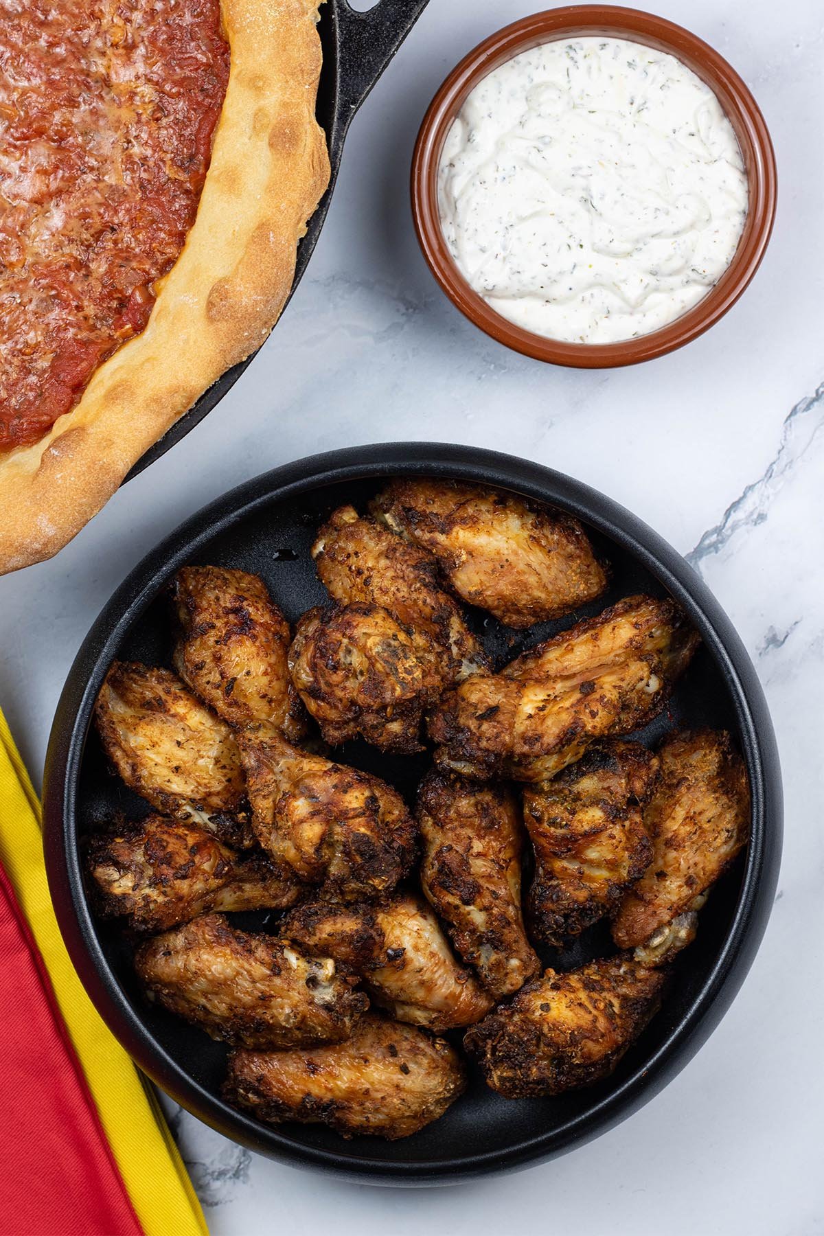 Easy air fryer Spanish chicken wings in a black serving bowl with a Chicago deep pan pizza and bowl of garlic and herb dip in the background.