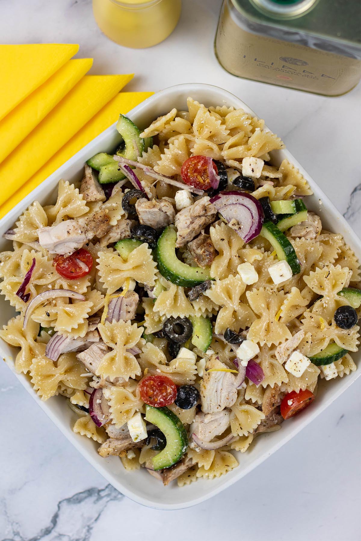 Greek chicken pasta salad in a square white serving dish with a yellow pepper grinder and tin of Greek extra virgin olive oil in the background.