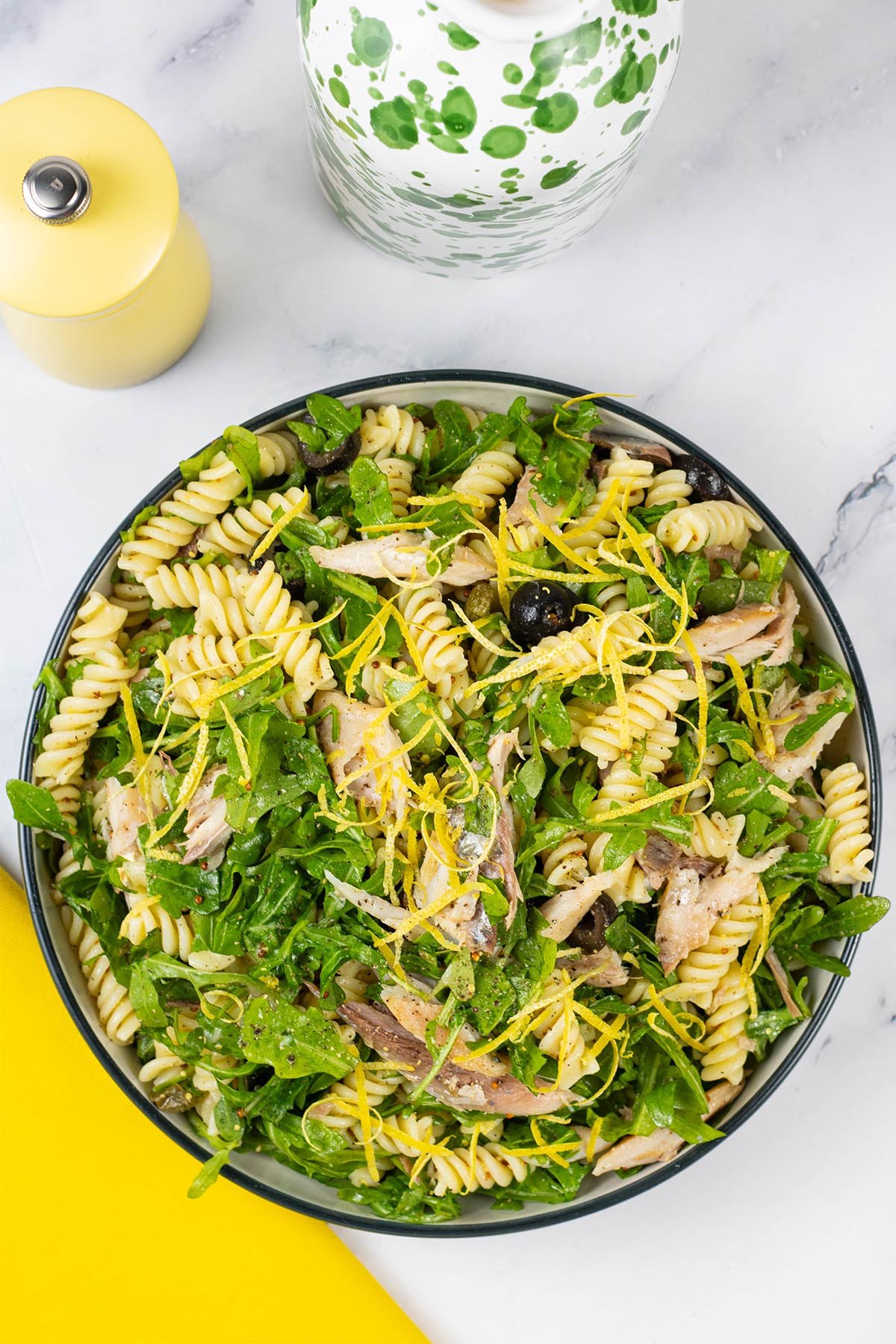 Hot smoked mackerel and rocket pasta salad in a black and cream serving bowl with a yellow pepper grinder and green and white oil bottle in the background.