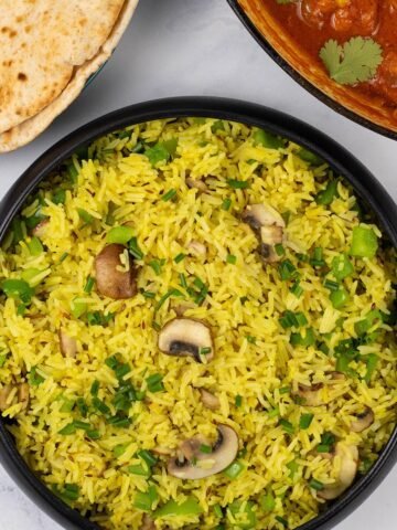 Mushroom rice in a black serving bowl with a plate of chapatis and dish of speedy gunpowder chicken in the background.