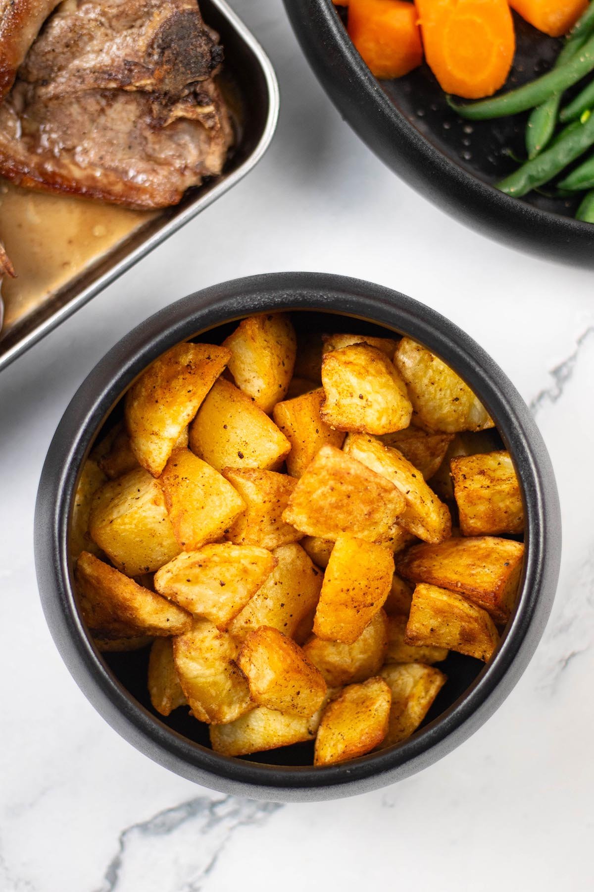 Paprika parmentier potatoes in a black serving bowl with pan-fried lamb chops and a bowl of carrots and green beans in the background.