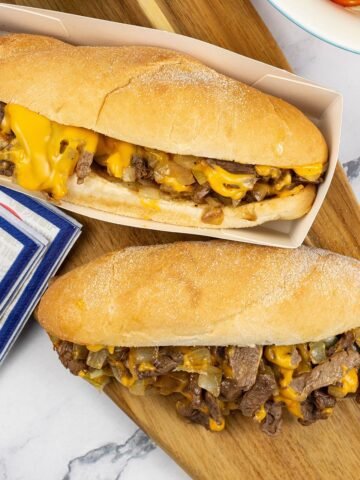 Two Philly cheesesteaks on a wooden board with red, white and blue napkins on the side.