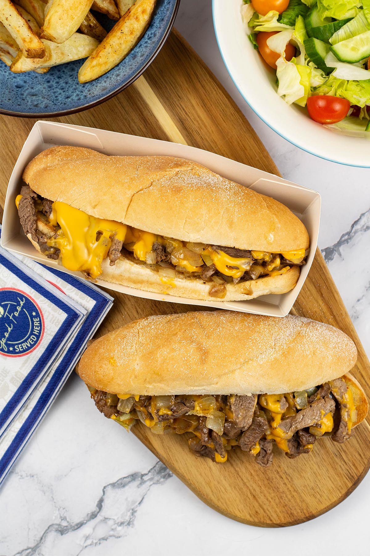 Two Philly cheesesteaks on a wooden board with a bowls of salad and chunky chips in the background.