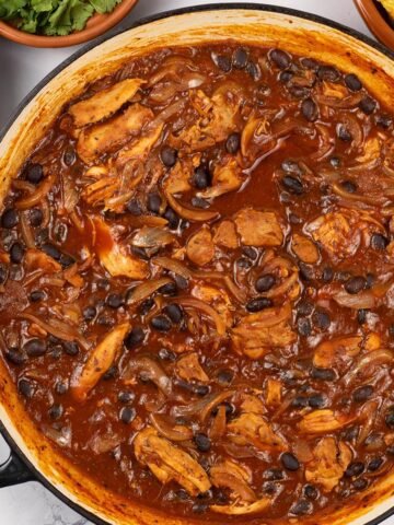 Pulled chicken and black bean chilli in a large round shallow casserole dish with dishes of rice, coriander and tortilla crisps on the side.
