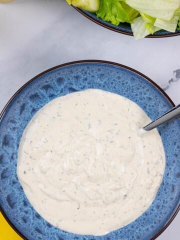 Ranch dressing in a blue patterned serving bowl with spoon and a bowl of lettuce salad in the background.