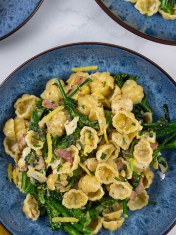 Two portions of smoked haddock with tenderstem broccoli, spinach and conchiglie in blue patterned bowls with a plate of crusty bread on the side.