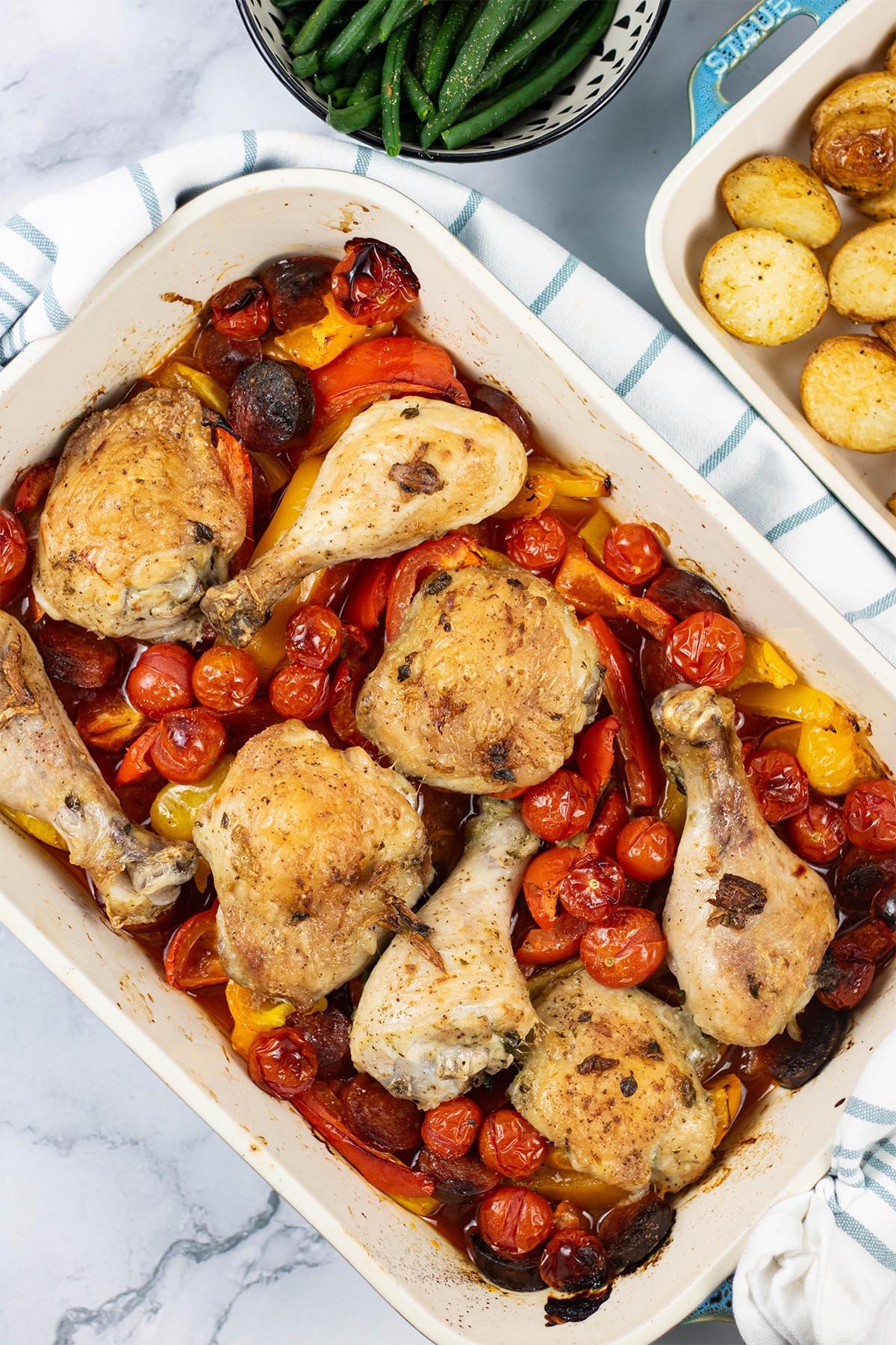 Spanish chicken tray bake in a large rectangle Staub oven dish with dishes of green beans and roast baby potatoes on the side.