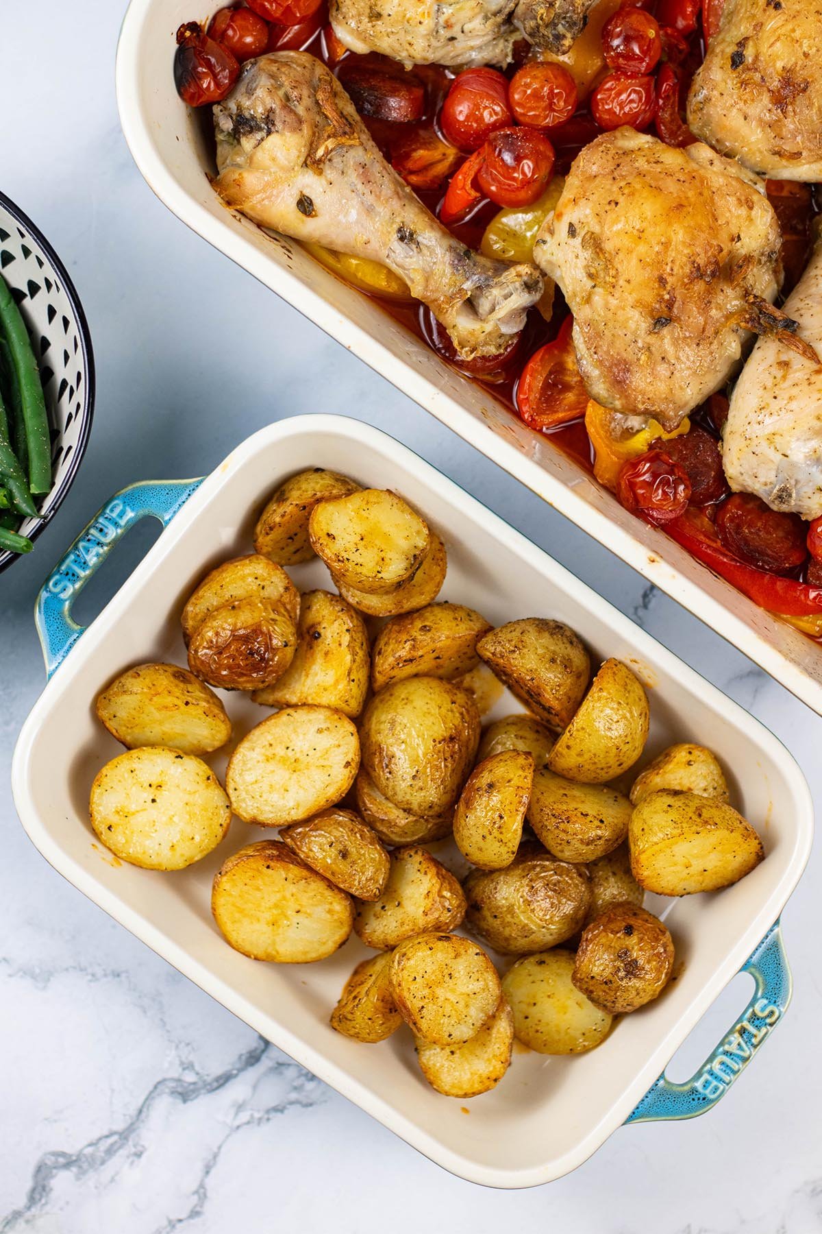 Spanish-style roast baby potatoes in a small rectangle Staub oven dish, with a large dish of Spanish chicken traybake in the background.