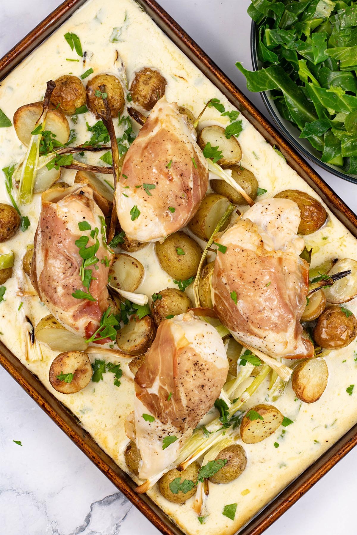 Four portions of parma ham, chicken and creamy potato traybake on a large baking tray, with a bowl of spring greens the side.