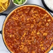 Slow cooked pork shoulder chilli in a large round casserole with bowls of nachos, spring onions and rice in the background.
