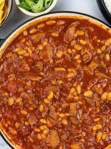 Slow cooked pork shoulder chilli in a large round casserole with bowls of nachos, spring onions and rice in the background.