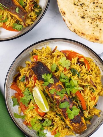 2 portions of Balti salmon rice bowls in black and white bowls with a wedge of lime and plate of garlic bread on the side.