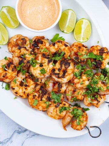Chilli and lime prawn skewers garnished with chopped coriander on a white oval platter with lime wedges and a dish of spicy mayo.