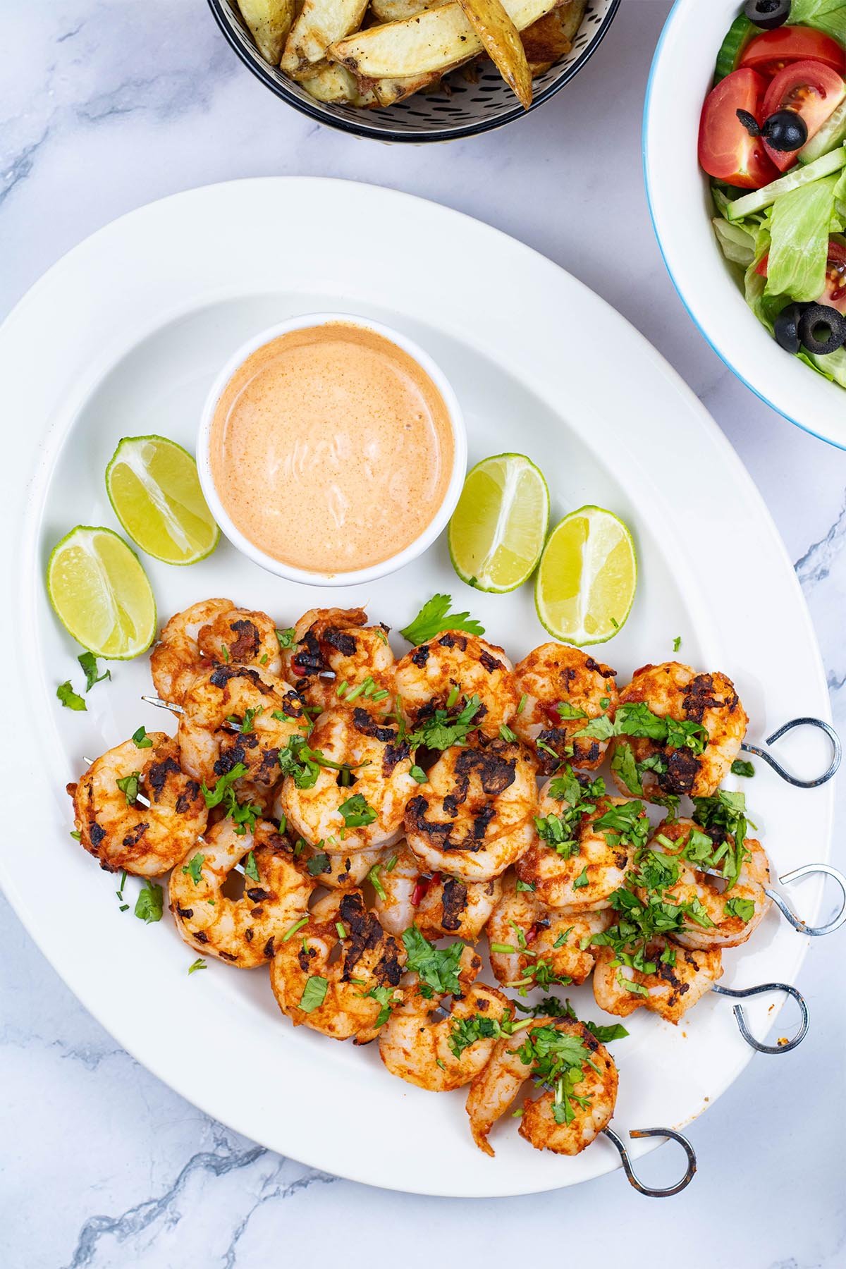 Chilli and lime prawn skewers garnished with chopped coriander on a white oval platter with lime wedges and a dish of spicy mayo.