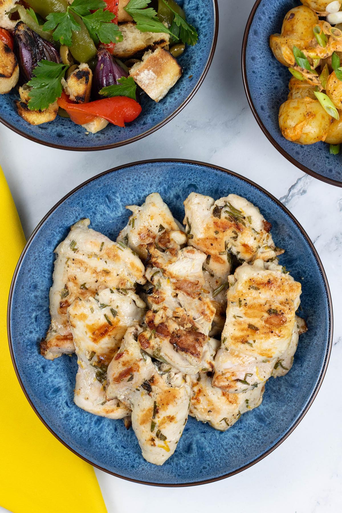 Lemon herb chicken thighs in a blue patterned serving bowl with a dish of barbecued mixed pepper salad on the side.