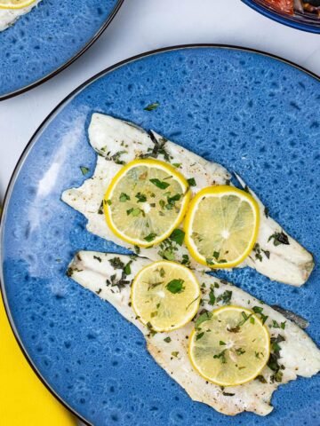2 x oregano and lemon barbecued sea bass fillets on a blue dinner plate with a bowl of Greek salad on the side.