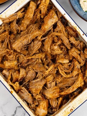 BBQ pulled pork in a blue rimmed white rectangle enamel roasting tin, with a bowl of coleslaw in the background.
