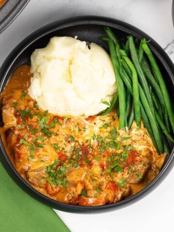 A portion of slow cooker beef goulash in a black bowl with mashed potatoes and green beans, and a green napkin on the side.