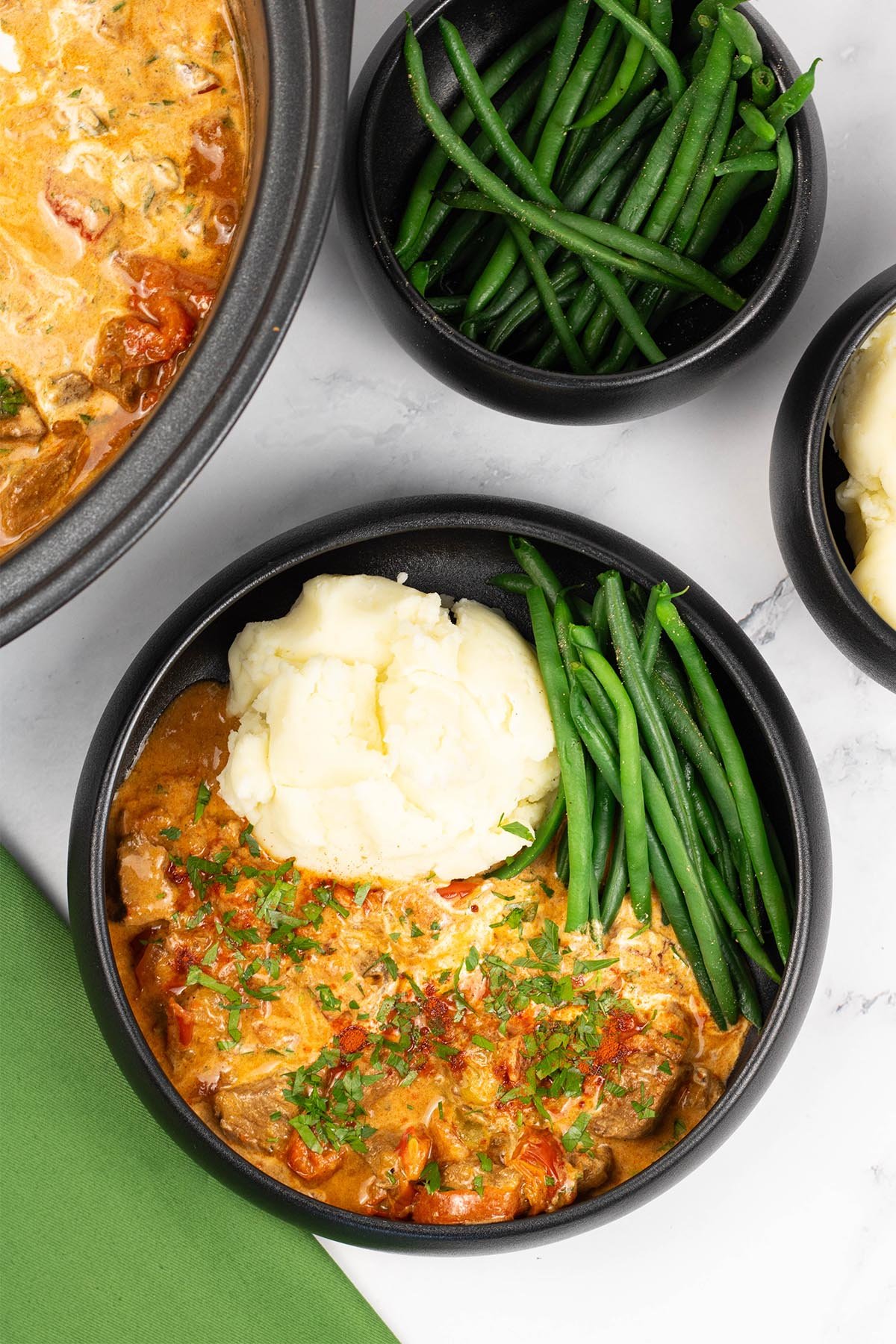 A portion of slow cooker beef goulash in a black bowl with mashed potatoes and green beans, and a green napkin on the side.