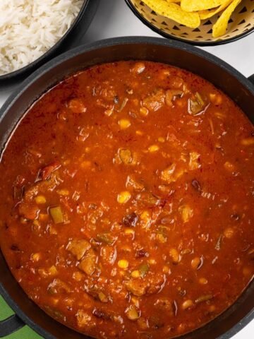 Smoky pork and beans made in the Thermomix in a black casserole pot, with serving bowls of rice and nachos in the background.