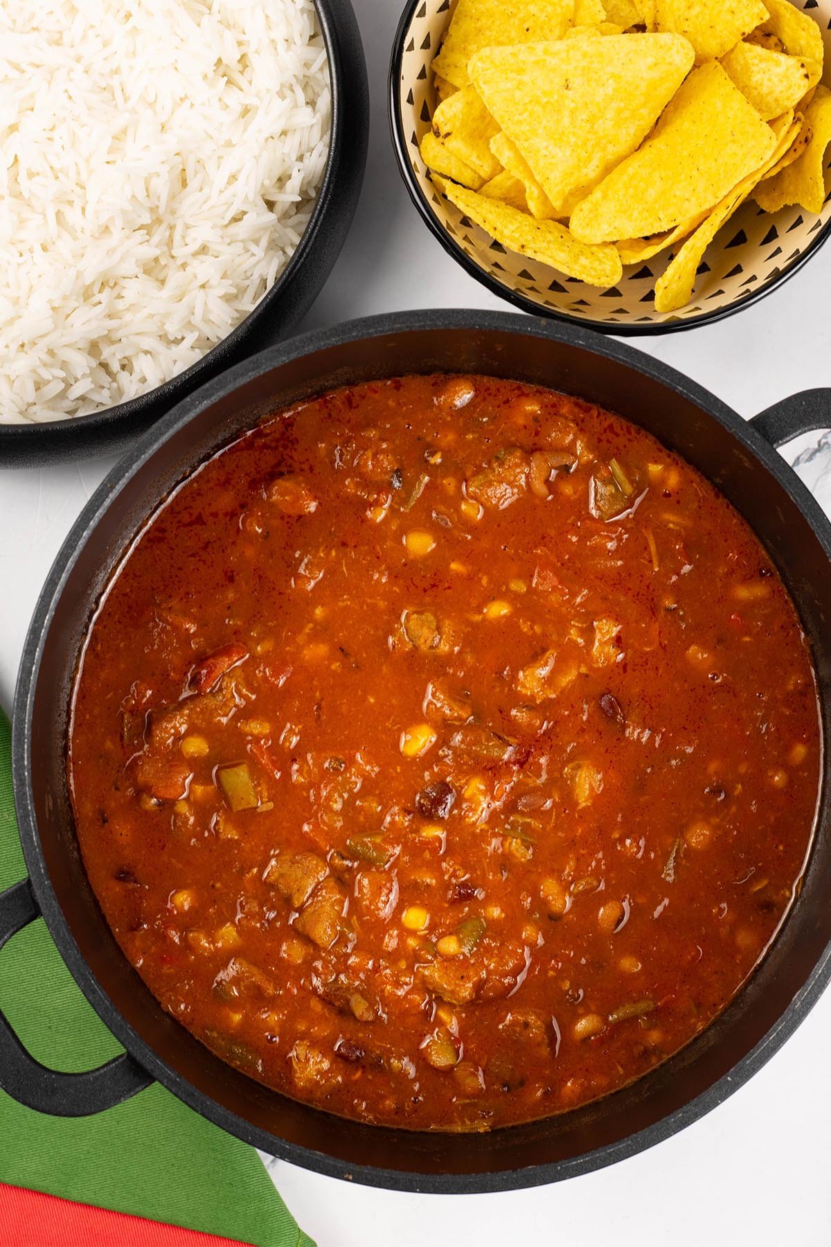 Smoky pork and beans made in the Thermomix in a black casserole pot, with serving bowls of rice and nachos in the background.