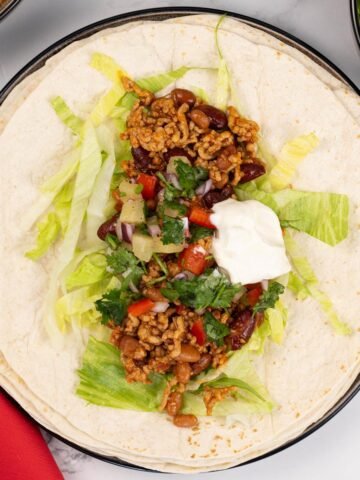 A smoky pork tortilla sitting on a stack of soft tortilla wraps, garnished with pineapple salsa and sour cream.