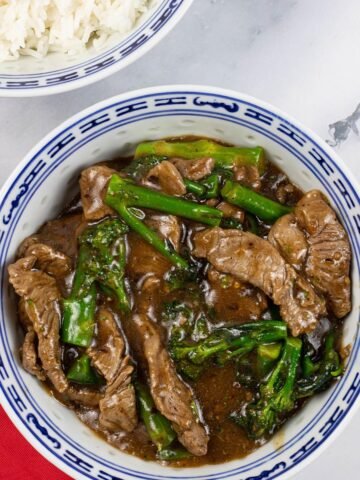 Spiced beef and broccoli in a blue and white oriental bowl with a bowl of steamed jasmine rice in the background.