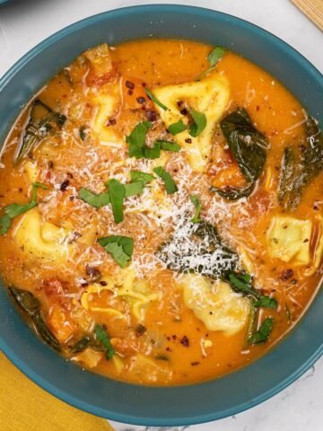 A portion of tomato and tortellini soup in a teal bowl with a mustard napkin on the side.