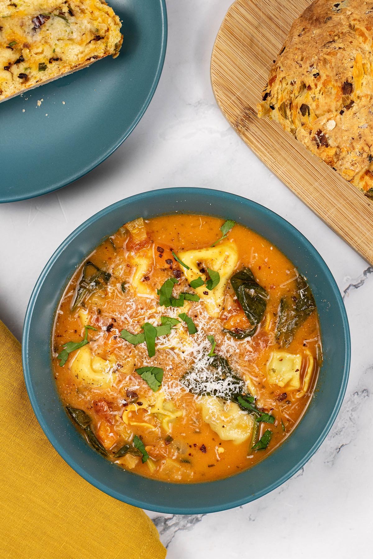 A portion of tomato and tortellini soup in a teal bowl with a plate of sliced leek and red leicester soda bread in the background.