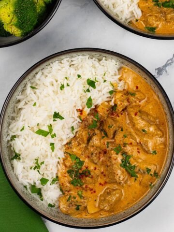 Two portions of slow cooker beef stroganoff in black and white bowls with rice and a bowl of green veg on the side.