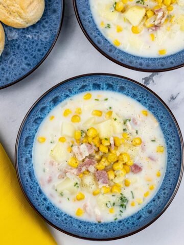 Two bowls of bacon and sweetcorn chowder in blue patterned bowls with a plate of crusty buns on the side.