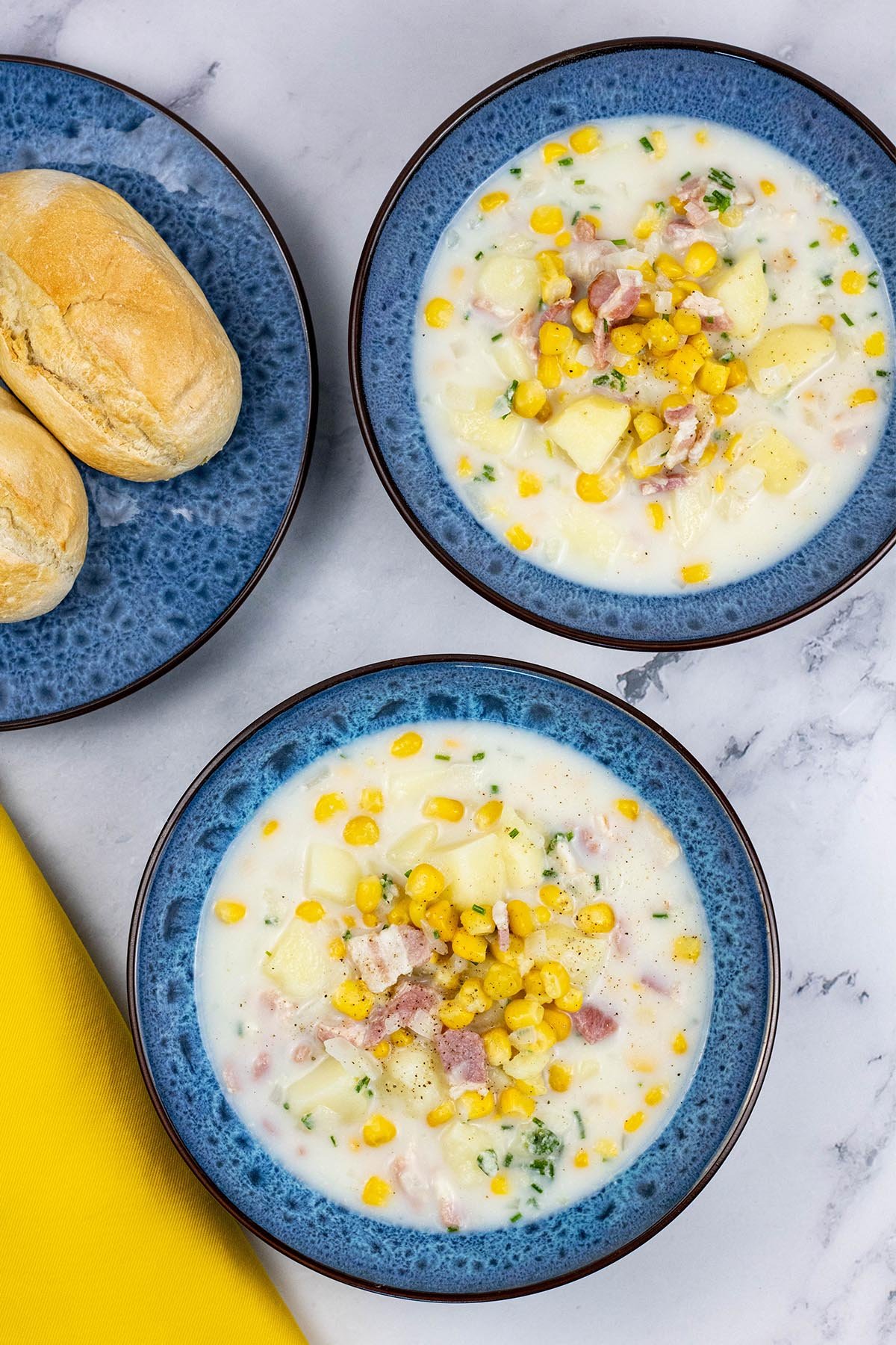 Two bowls of bacon and sweetcorn chowder in blue patterned bowls with a plate of crusty buns on the side.