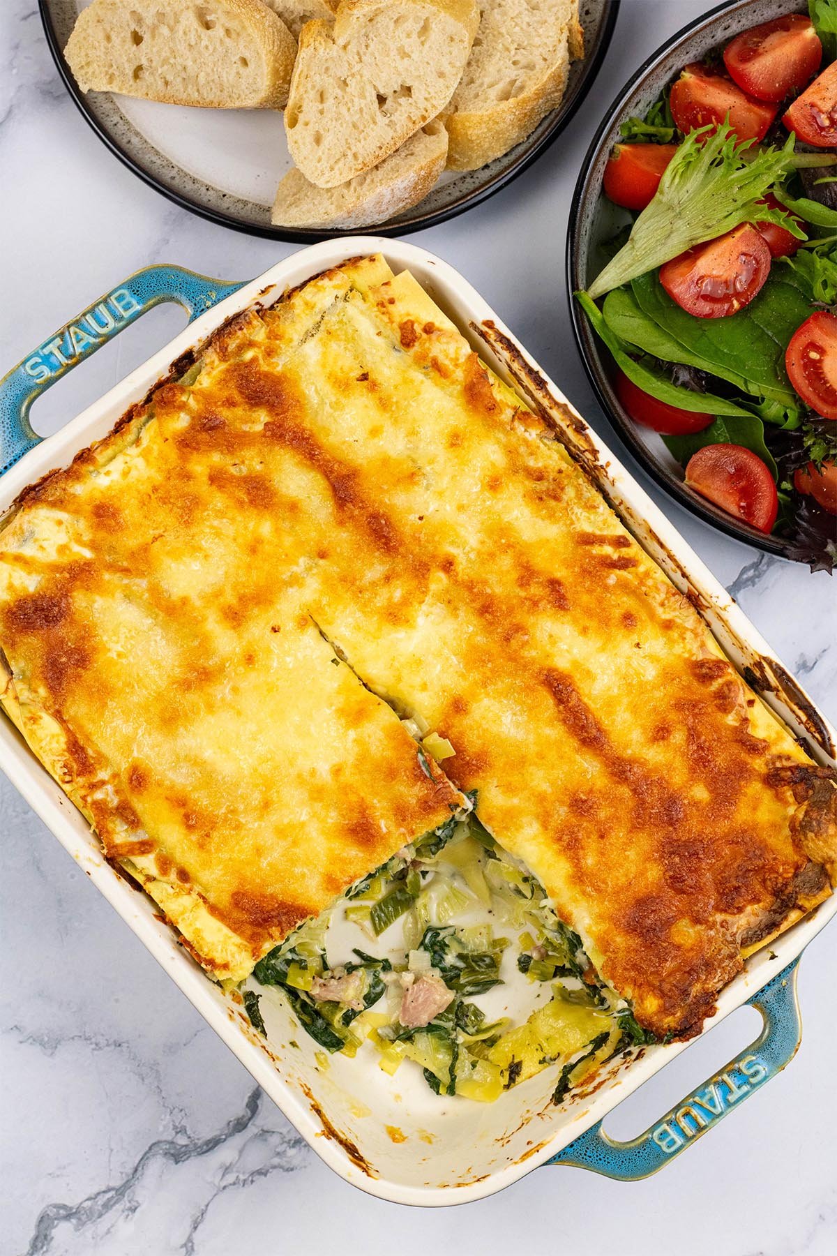 Ham, spinach and mozzarella lasagne in a rectangle Staub oven dish with a portion removed, a bowl of salad and plate of crusty bread on the side.