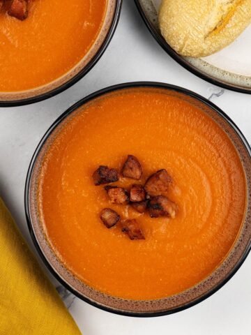 Two portions of red lentil and chorizo soup in black and white patterned bowls with a crusty baguette on a plate on the side.