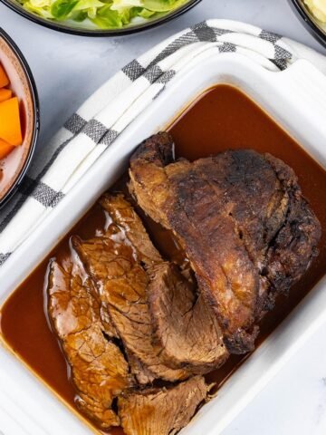 Slow cooker beef pot roast sitting in a white rectangular serving dish with gravy and a couple of slices carved.