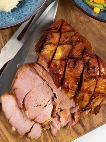 Slow cooker honey roast ham on a wooden board with two slices cut and bowls of mashed potatoes and warm carrot salad on the side.