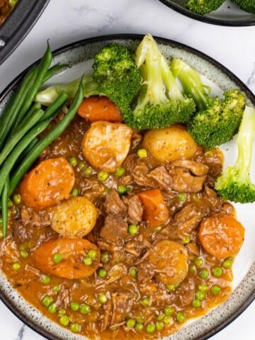 A portion of slow cooker old fashioned beef stew on a black and white patterned dinner plate with broccoli and green beans on the side.